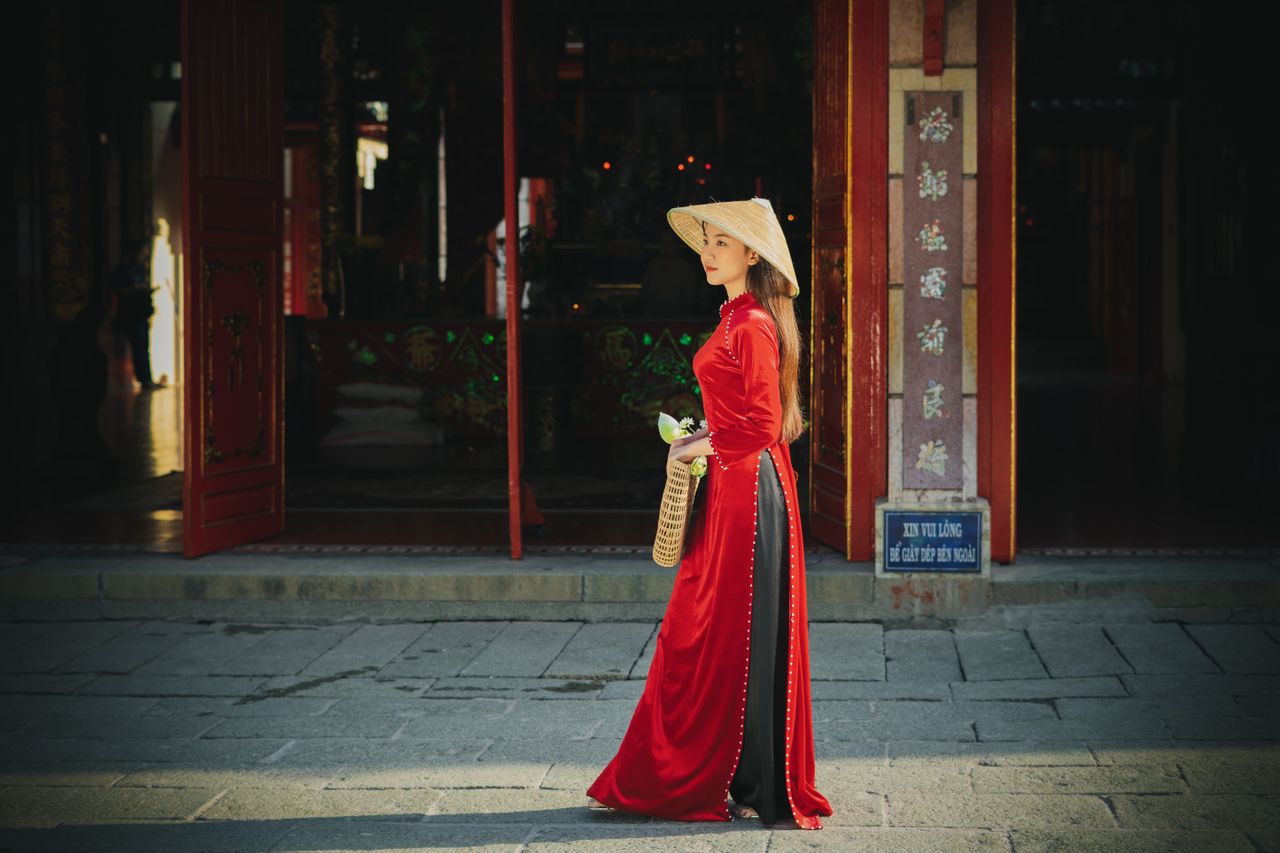 About An Asian woman in a red dress and conical hat walking outdoors, embodying cultural elegance.
