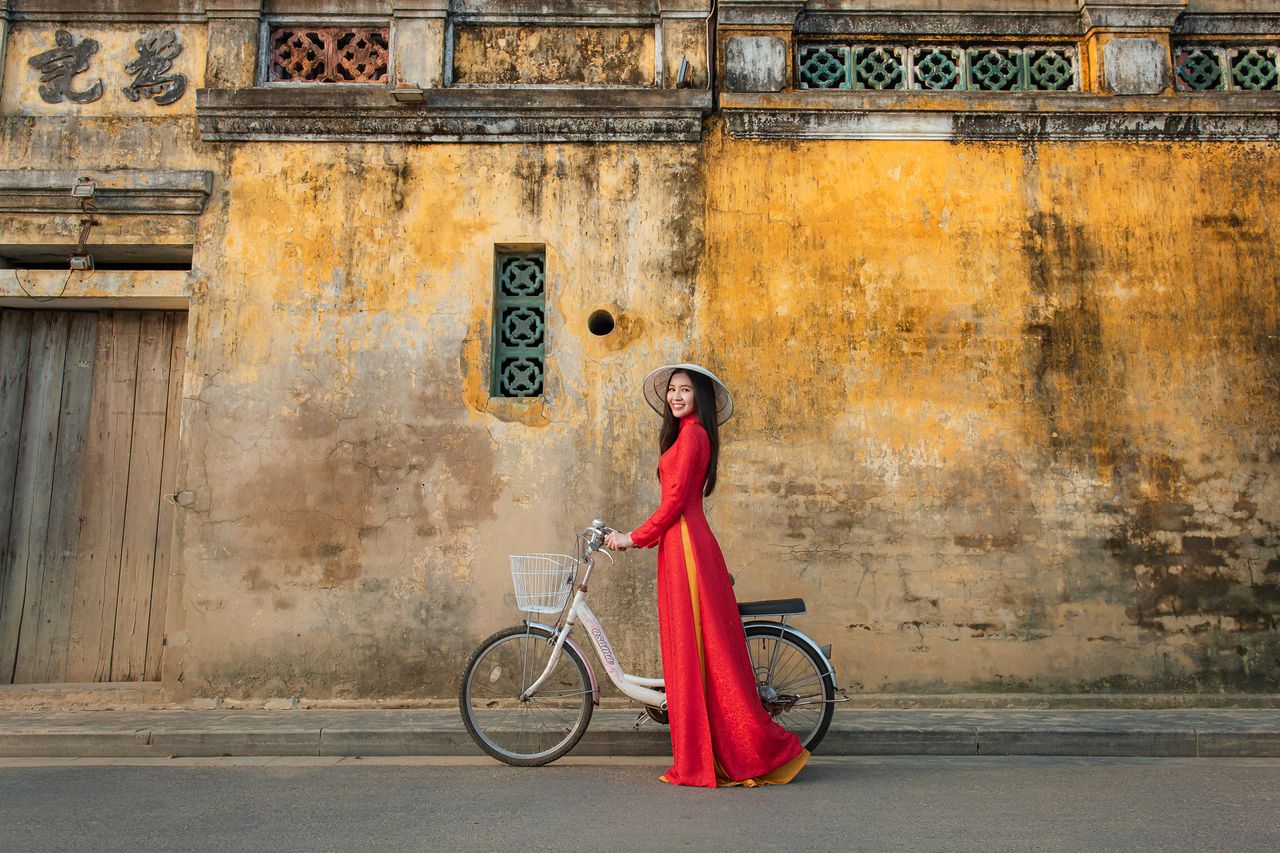 A woman in a red áo dài cycles past a historic wall in Hội An, Vietnam.
